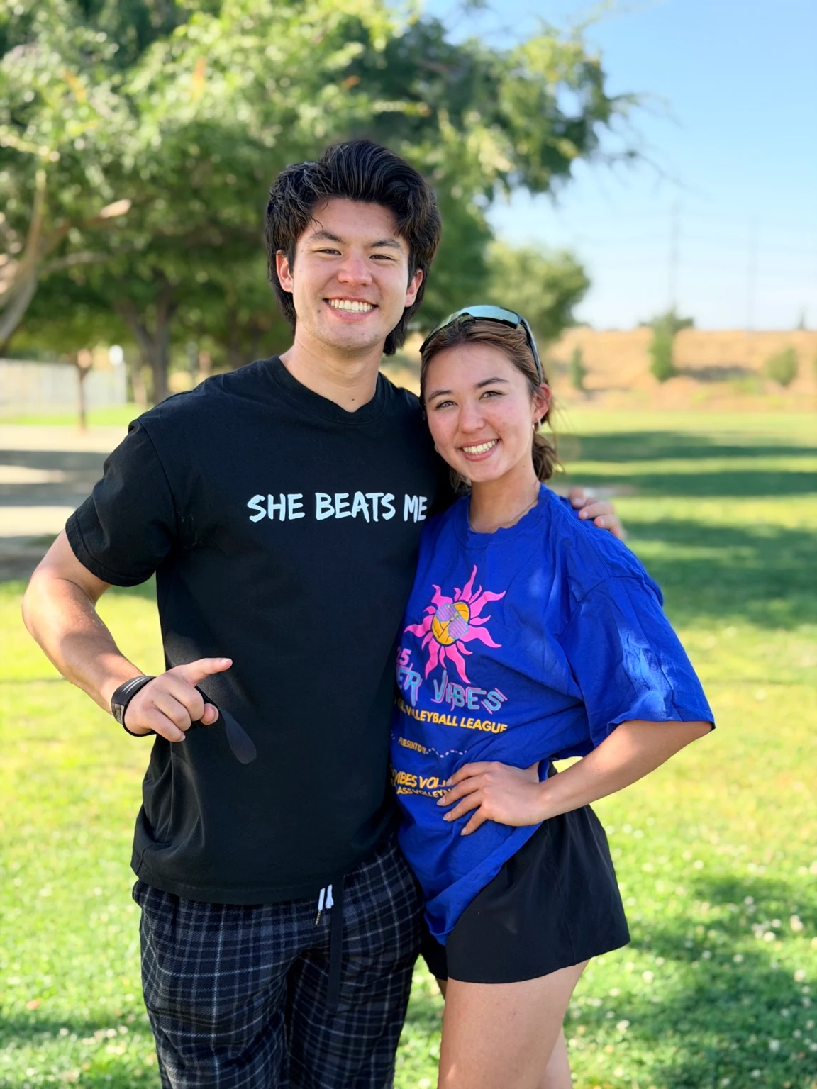 Two people smiling outdoors at a park. One wears the bright blue Summer Vibes league T-shirt with its pink sun and yellow volleyball graphic. Photograph taken and edited by DeAnthony Herbert.